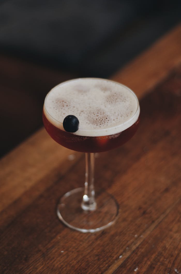A sophisticated cocktail served in a coupe glass, garnished with a cherry, on a wooden table.